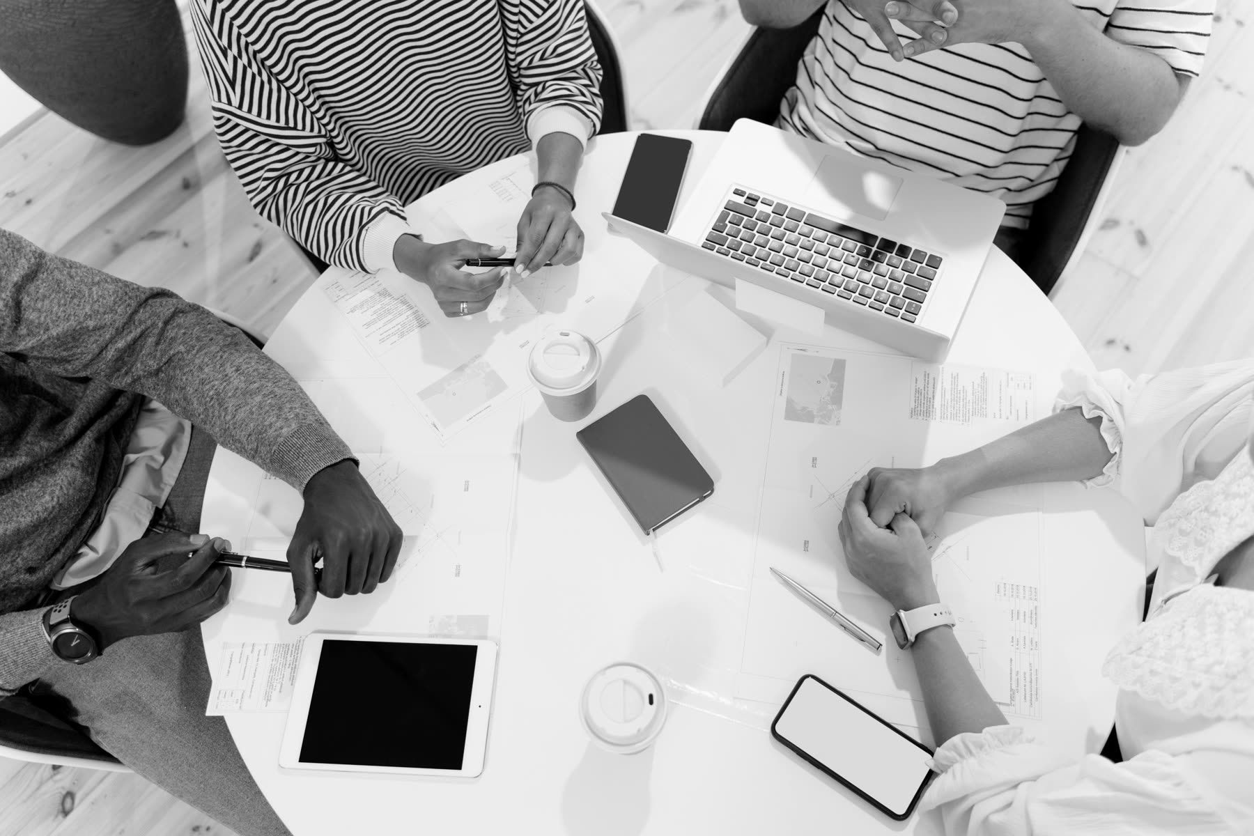 Overhead view of several people's hands around a round table with laptops, notebooks, and coffee during a working session