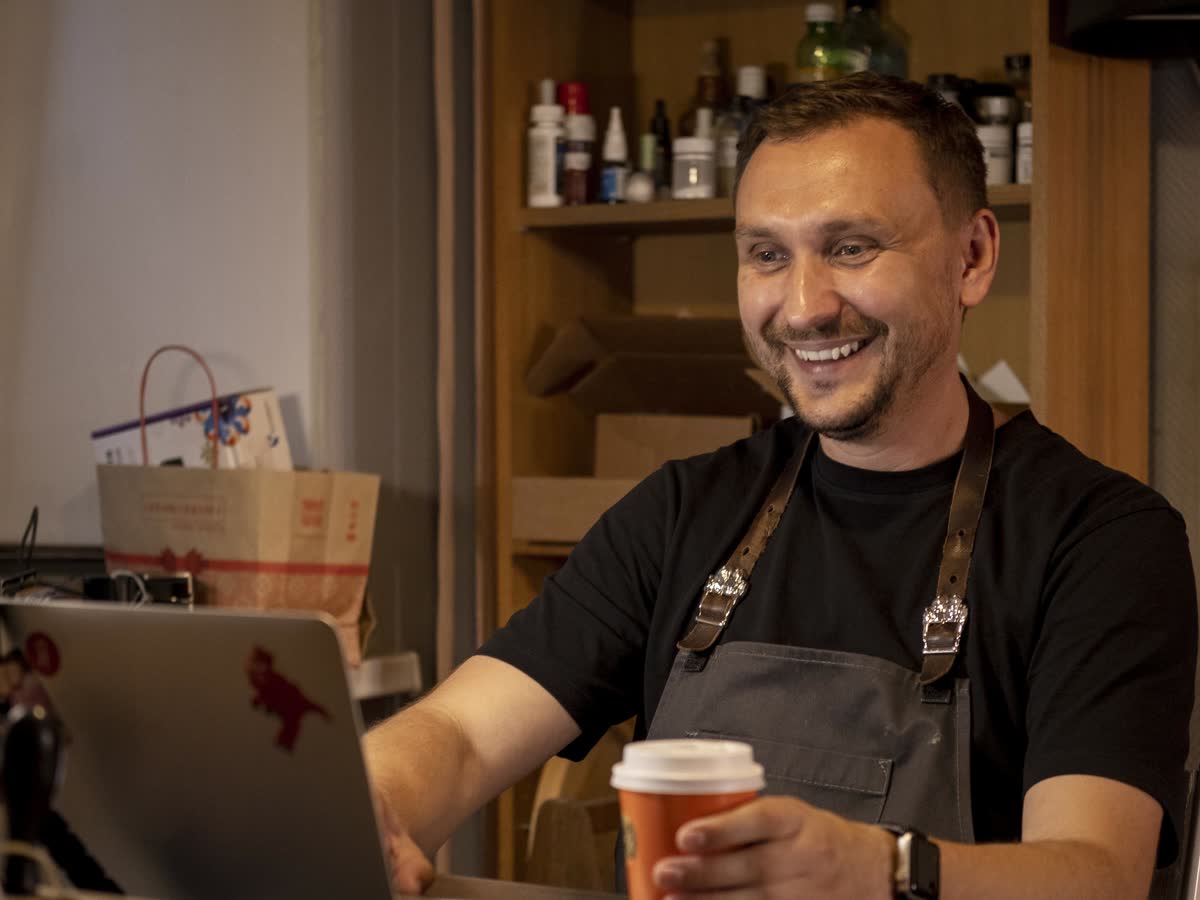 A smiling small-business owner in an apron works at his laptop in his shop